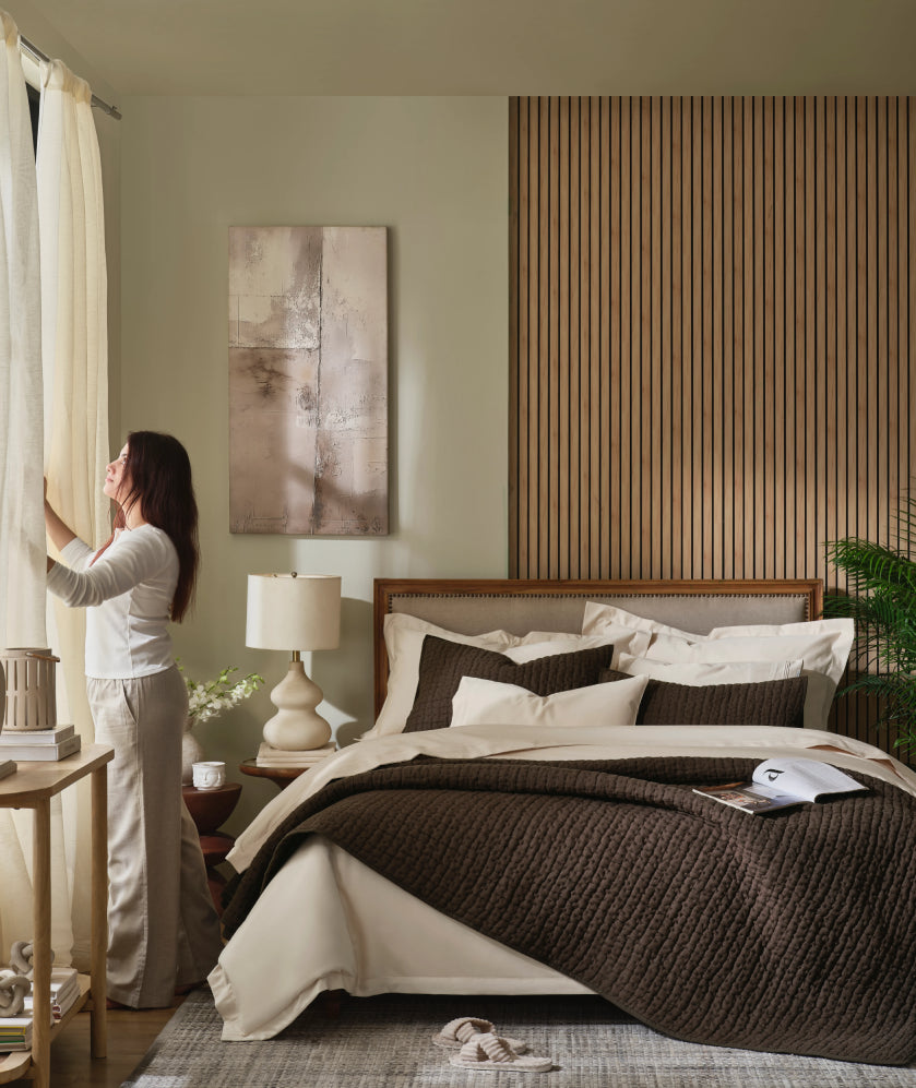 Woman opening curtains in a bedroom with a bed dressed in an espresso quilt and cream linen