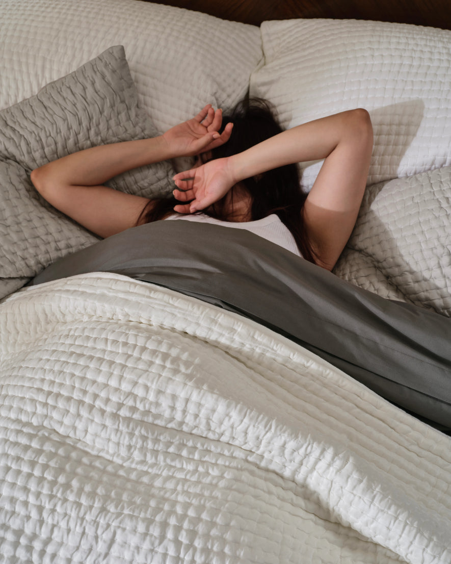 Person stretching awake in bed under a white quilt and grey sheets