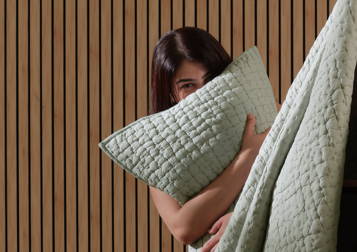 Woman hugging a sage green quilted pillow and quilt against a wood-slat wall